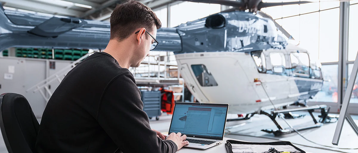 Mitarbeiter der DRF Aviation Services arbeitet an einem Laptop vor einem Hubschrauber in einem Hangar.
