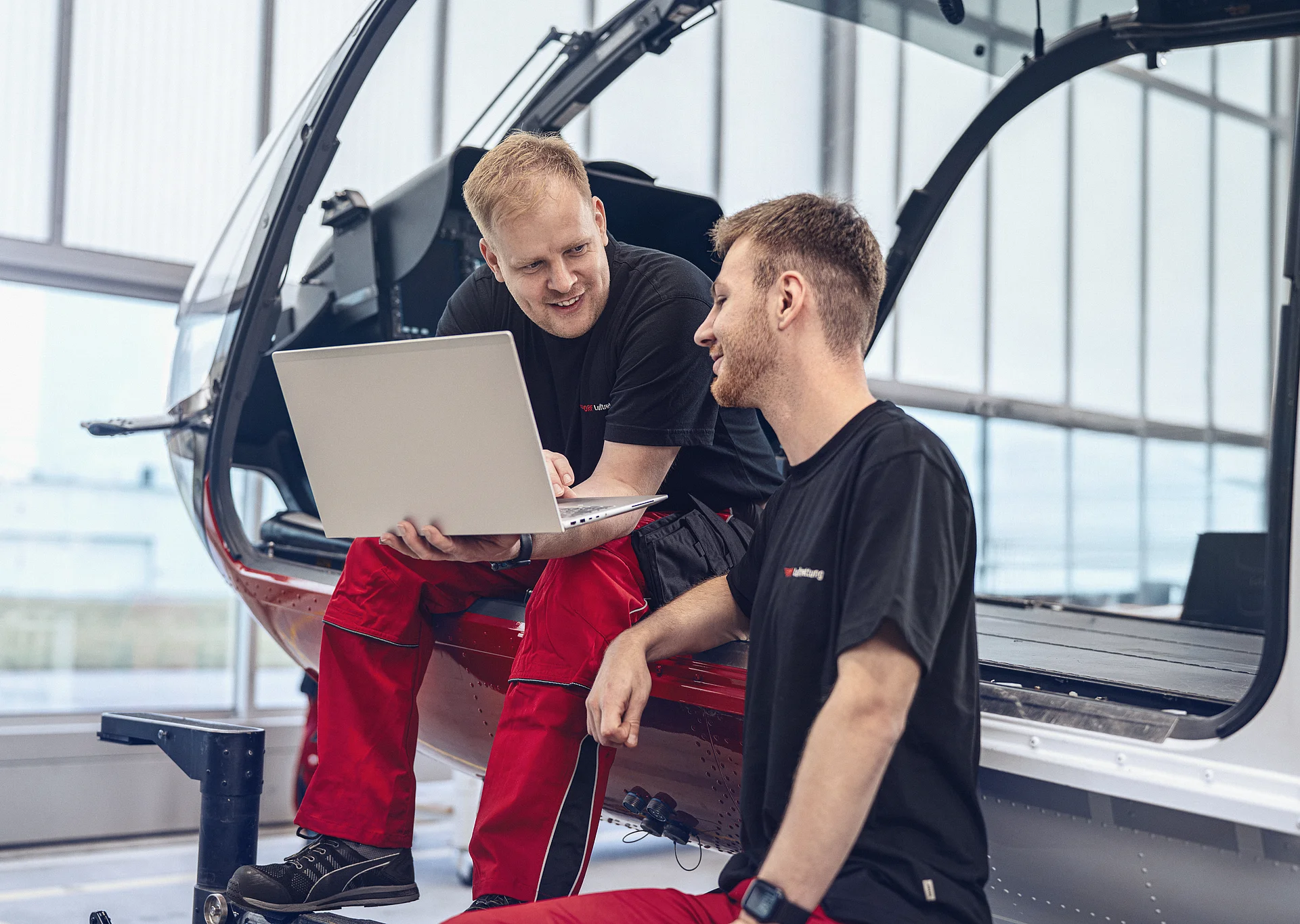 Zwei Techniker der DRF Luftrettung blicken auf einen Laptopbildschirm und sitzen dabei auf der Kufe eines Hubschraubers.